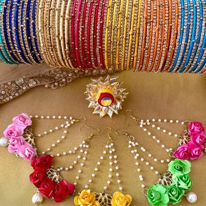 May include: A collection of colorful bangles in shades of blue, gold, red, pink, orange, and yellow, adorned with small, sparkling embellishments. Below are floral earrings with pearl accents and a decorative flower.