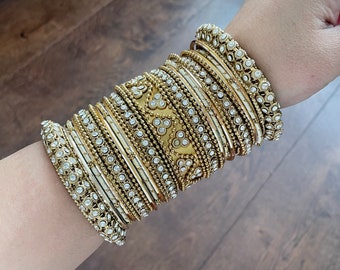 Op de afbeelding: Een set gouden armbanden met witte strass steentjes. De armbanden zijn op elkaar gestapeld en hebben een decoratief ontwerp.