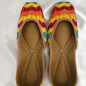 K&ouml;nnte beinhalten: Ein Paar flache, bestickte Schuhe mit einem gelben, orangefarbenen, roten und blauen Muster. Die Schuhe haben eine braune Ledersohle und eine spitze Zehenpartie.