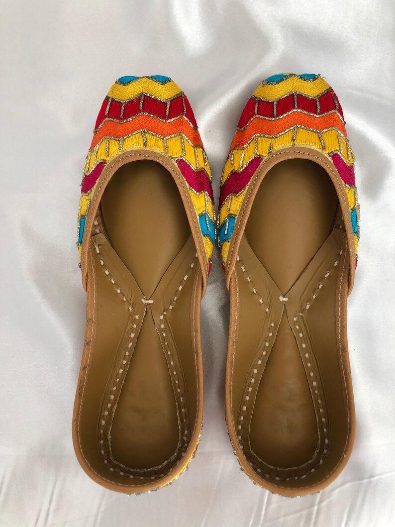 K&ouml;nnte beinhalten: Ein Paar flache, bestickte Schuhe mit einem gelben, orangefarbenen, roten und blauen Muster. Die Schuhe haben eine braune Ledersohle und eine spitze Zehenpartie.