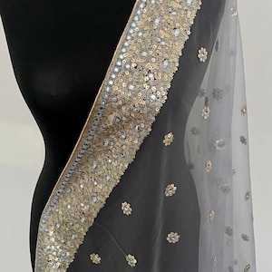 May include: A sheer, pale grey scarf with an ornate border. The border is adorned with gold sequins, silver details, and small floral patterns. The scarf is draped over a black mannequin.