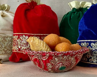 May include: A red, green, and blue velvet pouch set with gold embroidery. The pouches are filled with small, round, orange sweets and a small, golden pastry. The pouches are sitting on a brown, textured surface.