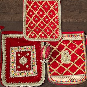 Puede incluir: Tres bolsas de terciopelo rojo con intrincados bordados y adornos dorados. Las bolsas presentan patrones geométricos y florales, con una variedad de acentos coloridos. Una bolsa tiene un diseño de cachemira y otra un diseño floral central.