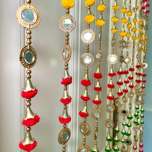 May include: Decorative beaded door hanging with gold-colored accents, mirrors, and colorful pom-poms. The hanging features yellow, red, green, and pink elements. The design includes round mirrors and bell-shaped ornaments.