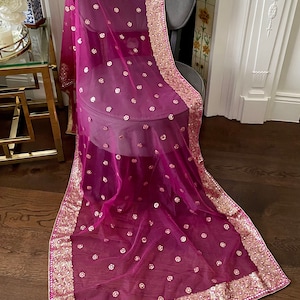 May include: A sheer, magenta dupatta, or scarf, is draped over a grey chair. The scarf is decorated with small, embroidered floral accents and a wide, gold sequined border. The fabric is lightweight and appears delicate.