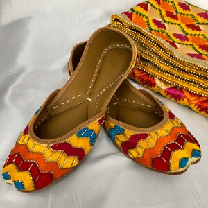 K&ouml;nnte beinhalten: Ein Paar bunte, bestickte flache Schuhe mit einem geometrischen Muster in Gelb, Rot, Orange, Blau und Wei&szlig;. Die Schuhe sind aus Leder und haben eine spitze Zehenpartie.