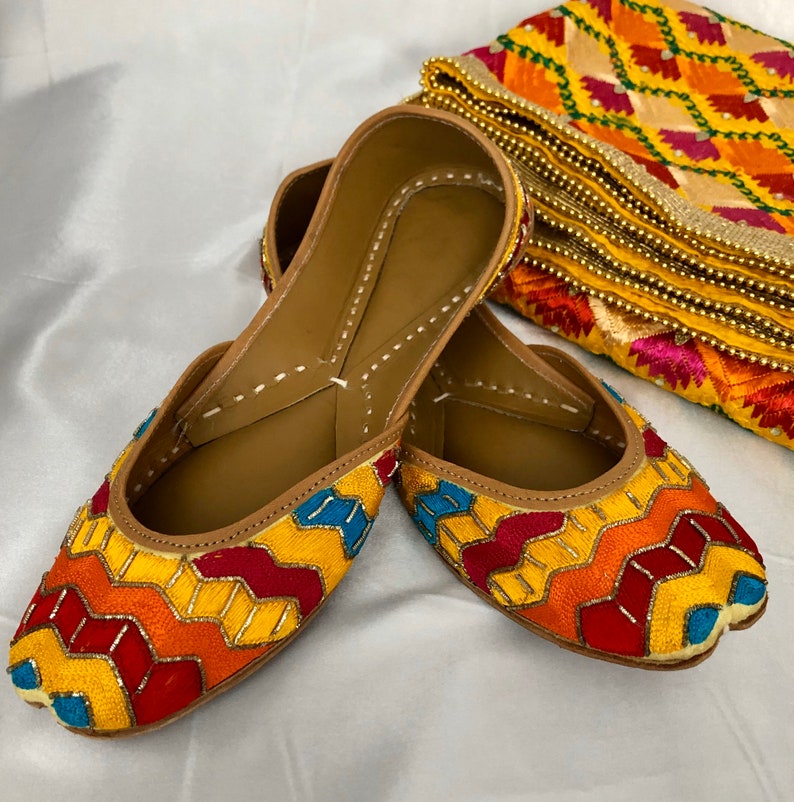 K&ouml;nnte beinhalten: Ein Paar bunte, bestickte flache Schuhe mit einem geometrischen Muster in Gelb, Rot, Orange, Blau und Wei&szlig;. Die Schuhe sind aus Leder und haben eine spitze Zehenpartie.