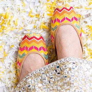 K&ouml;nnte beinhalten: Ein Paar flache Schuhe mit bunten, gestickten Chevron-Mustern. Die Schuhe sind hellbraun mit einem pinkfarbenen, gelben, orangen, roten und blauen Chevron-Muster. Die Schuhe befinden sich auf einem wei&szlig;en Hintergrund.