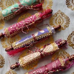 Può includere: Vista dall'alto di cinque oggetti decorativi a forma di pergamena. Ogni pergamena è avvolta in tessuto fantasia in vari colori, tra cui verde, rosa, viola e crema. Sono adornati con finiture dorate, nappe rosse e legati con un nastro abbinato.