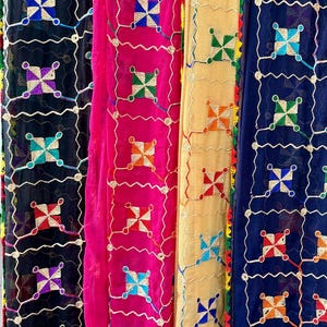 Peut inclure: Gros plan de plusieurs écharpes brodées colorées. Les écharpes sont dans des tons de noir, rose, or et bleu. Chaque écharpe présente un motif carré répétitif avec des motifs de point de croix colorés. Les bords sont garnis de coutures décoratives.