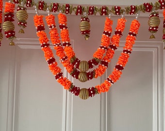 May include: A traditional Indian door hanging garland made of orange and red fabric flowers with gold and white accents. The garland is decorated with small, round, gold ornaments.