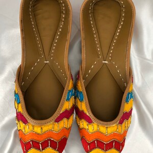 K&ouml;nnte beinhalten: Ein Paar flache, bestickte Schuhe mit einem Zickzackmuster in Gelb-, Orange-, Rot- und Blaut&ouml;nen. Die Schuhe haben eine braune Ledersohle und eine spitze Zehenpartie.