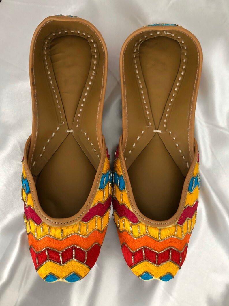 K&ouml;nnte beinhalten: Ein Paar flache, bestickte Schuhe mit einem Zickzackmuster in Gelb-, Orange-, Rot- und Blaut&ouml;nen. Die Schuhe haben eine braune Ledersohle und eine spitze Zehenpartie.