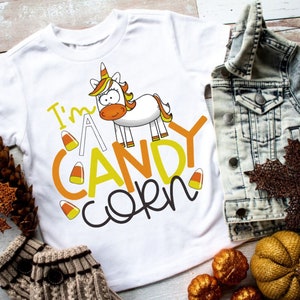 Może przedstawiać: Biała koszulka z kreskówkowym jednorożcem i napisem "I'm a Candy Corn". Koszulka jest otoczona jesiennymi ozdobami, w tym dżinsową kurtką, brązowym liściem i małymi dyniami.