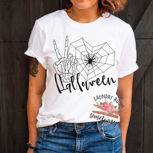 Puede incluir: Camiseta blanca con un gráfico en blanco y negro de una mano de esqueleto haciendo un signo de paz y una telaraña con una araña en el centro. El texto "Halloween" está escrito debajo del gráfico. La camiseta tiene un pequeño logotipo en la esquina inferior derecha que dice "Laundry Room Sanctuary".
