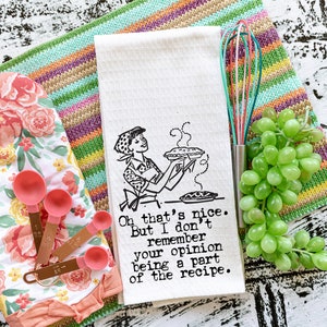 May include: A white tea towel with a black and white illustration of a woman holding a pie. The text on the towel reads "Oh that's nice. But I don't remember your opinion being a part of the recipe."