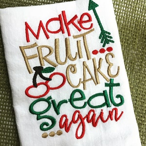 Puede incluir: Toalla de cocina blanca con bordado rojo, verde y dorado que dice "Make Fruit Cake Great Again" con un diseño de cereza roja y hoja verde.