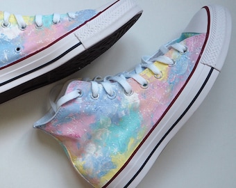 May include: A pair of high-top Converse sneakers with a tie-dye design in shades of pink, blue, and yellow. The sneakers have a white rubber sole and a black stripe around the edge.