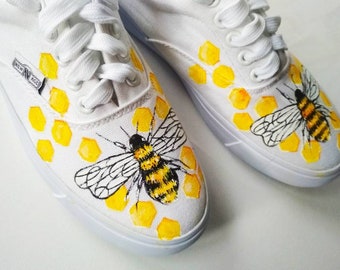 May include: White canvas shoes with a hand-painted design of a bee and honeycomb. The bee is yellow and black with transparent wings. The honeycomb is yellow.