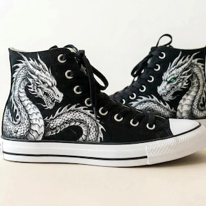 Könnte beinhalten: Schwarze High-Top-Sneakers mit weißen Gummisohlen. Jeder Schuh hat ein handgemaltes Drachendesign in Weiß und Grau. Die Augen des Drachen sind grün. Die Sneakers haben schwarze Schnürsenkel und silberne Ösen.