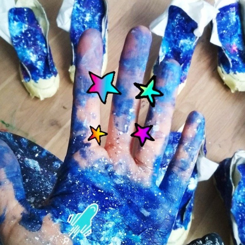 May include: A hand painted with blue and white galaxy-themed paint with colorful star stickers and a rocket sticker. The hand is surrounded by galaxy-themed sneakers.