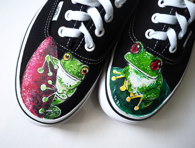 May include: A pair of black canvas shoes with white laces, painted with green frogs. One frog is on a pink background and the other is on a green background. The shoes have the word "VANS" printed on the side.