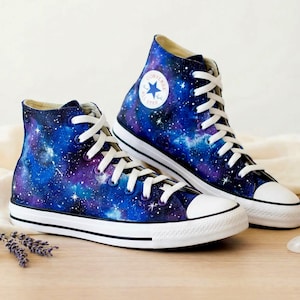 Op de afbeelding: Een paar hoge sneakers met een levendige sterrenstelselprint in tinten diepblauw, paars en zwart met witte steraccenten. De schoenen hebben witte veters, witte rubberen neuzen en een witte zool met een zwarte streep. Een ronde Converse All Star-patch is zichtbaar aan de zijkant van de enkel.