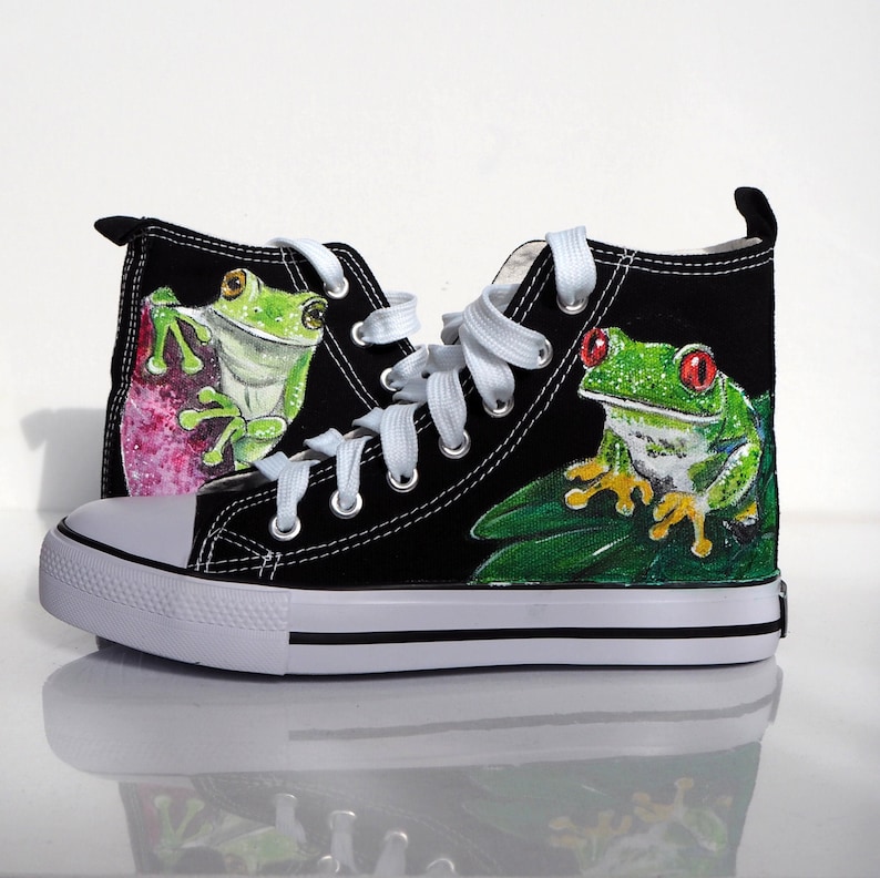 May include: Black canvas high-top sneakers with a hand-painted design of two green tree frogs. One frog is on a pink flower and the other is on a green leaf.