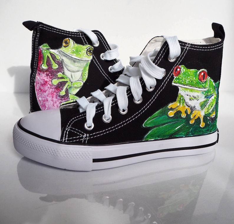 May include: Black canvas high top sneakers with white laces. One sneaker has a green frog painted on it with red eyes and yellow toes. The other sneaker has a green frog painted on it with a pink background.