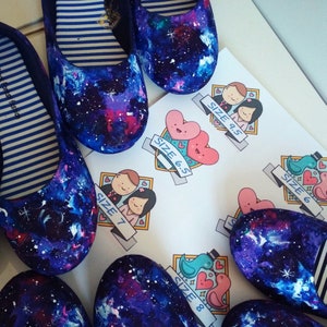 May include: A pair of galaxy-print canvas shoes with a white stripe on the side. The shoes are displayed with tags indicating sizes 6.5, 7, 8, 9.5.