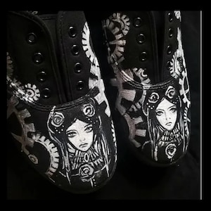 May include: A pair of black canvas shoes with white painted designs. The shoes feature a steampunk theme with gears and a woman's face.