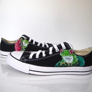 May include: Black canvas low-top sneakers with a hand-painted design of green frogs with pink eyes. The frogs are sitting on lily pads.