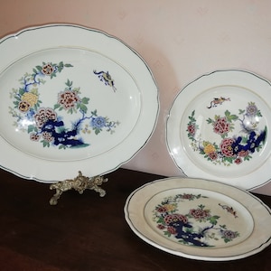May include: Three white ceramic plates with a floral design in shades of blue, red, yellow, and green. The plates are arranged in a pyramid shape, with the largest plate at the bottom and the smallest plate at the top.
