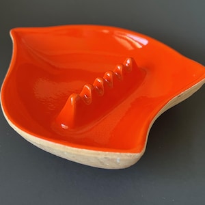 May include: A bright orange ceramic ashtray shaped like a leaf with a smooth, glossy interior finish. The center features a raised ridge with five distinct notches for resting items. The exterior edge shows a natural, unglazed beige clay texture.