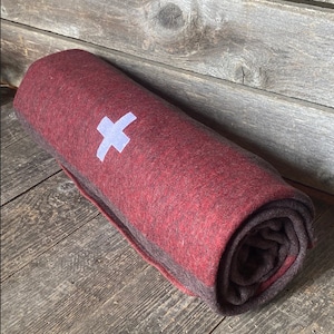 May include: A rolled up red and brown wool blanket with a white cross embroidered on it.