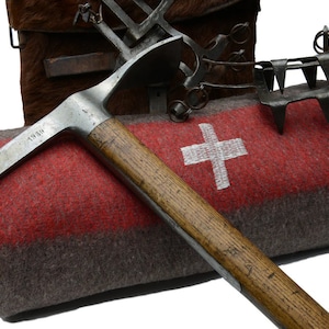 May include: A vintage ice axe with a wooden handle and a metal head, marked "1938", resting on a rolled-up red and gray blanket with a white Swiss cross.