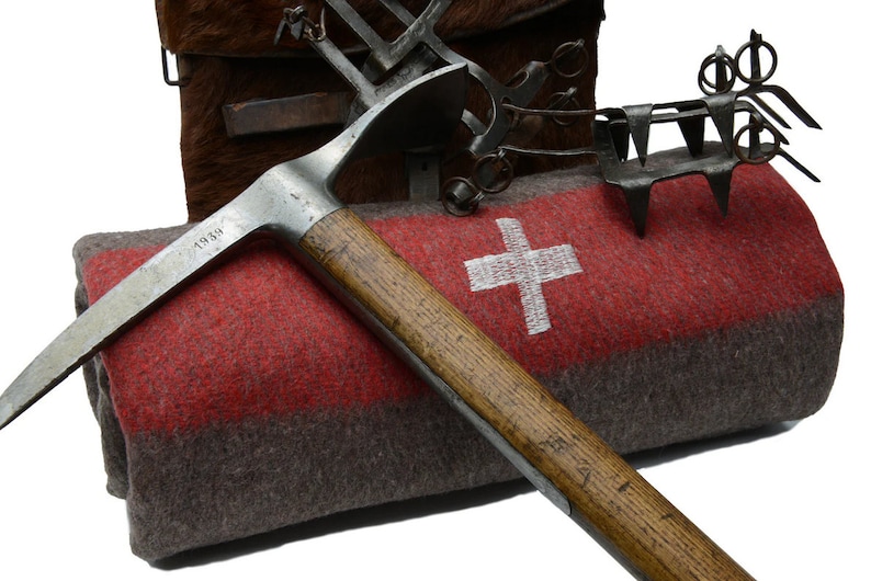 May include: A vintage ice axe with a wooden handle and a metal head, marked "1938", resting on a rolled-up red and gray blanket with a white Swiss cross.