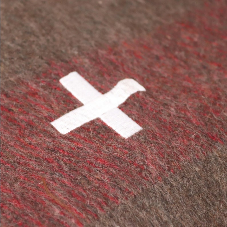 May include: A close-up of a red and gray wool blanket with a white cross in the center. The blanket is textured and has a fuzzy appearance.