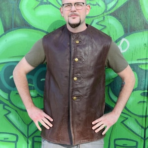May include: A brown leather vest with four gold buttons. The vest is sleeveless and has a round collar. The person is wearing a green t-shirt underneath. The background is a green mural.