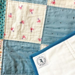 K&ouml;nnte beinhalten: Eine blaue und wei&szlig;e Patchwork-Decke mit wei&szlig;er R&uuml;ckseite und blauem Rand. Die Decke hat ein kleines Etikett, das in die Ecke eingen&auml;ht ist und "bits o' everything inc. little" lautet.