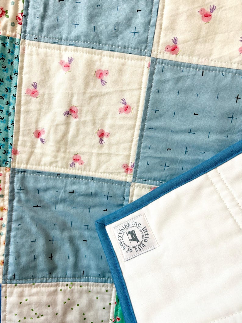K&ouml;nnte beinhalten: Eine blaue und wei&szlig;e Patchwork-Decke mit wei&szlig;er R&uuml;ckseite und blauem Rand. Die Decke hat ein kleines Etikett, das in die Ecke eingen&auml;ht ist und "bits o' everything inc. little" lautet.
