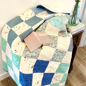 K&ouml;nnte beinhalten: Eine Patchwork-Decke in den Farben Blau, Wei&szlig; und Gr&uuml;n. Die Decke ist gefaltet und &uuml;ber einen Holztisch mit einem Buch darauf gelegt. Eine Vase mit Blumen steht auf dem Tisch.