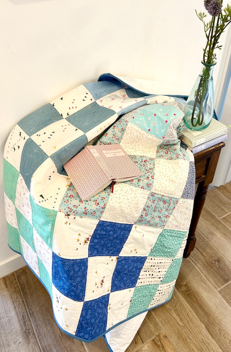 K&ouml;nnte beinhalten: Eine Patchwork-Decke in den Farben Blau, Wei&szlig; und Gr&uuml;n. Die Decke ist gefaltet und &uuml;ber einen Holztisch mit einem Buch darauf gelegt. Eine Vase mit Blumen steht auf dem Tisch.