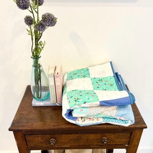 K&ouml;nnte beinhalten: Eine gefaltete blaue und gr&uuml;ne Patchwork-Decke mit wei&szlig;em Hintergrund und Blumenmuster liegt auf einem Holztisch mit Schublade. Eine Vase mit lila Blumen steht auf dem Tisch neben drei B&uuml;chern.