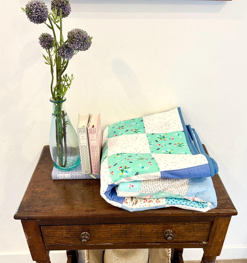 K&ouml;nnte beinhalten: Eine gefaltete blaue und gr&uuml;ne Patchwork-Decke mit wei&szlig;em Hintergrund und Blumenmuster liegt auf einem Holztisch mit Schublade. Eine Vase mit lila Blumen steht auf dem Tisch neben drei B&uuml;chern.