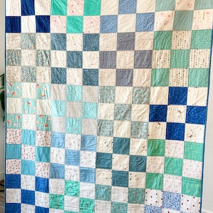 K&ouml;nnte beinhalten: Eine Patchwork-Decke mit einem Schachbrettmuster in Blaut&ouml;nen, Gr&uuml;n- und Wei&szlig;t&ouml;nen. Die Decke besteht aus verschiedenen Stoffen mit Blumen- und geometrischen Mustern.