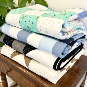 K&ouml;nnte beinhalten: Ein Stapel von vier gefalteten Quilts in verschiedenen Mustern und Farben, darunter Blau, Wei&szlig;, Gr&uuml;n und Schwarz, auf einem Holztisch mit einer Schublade.