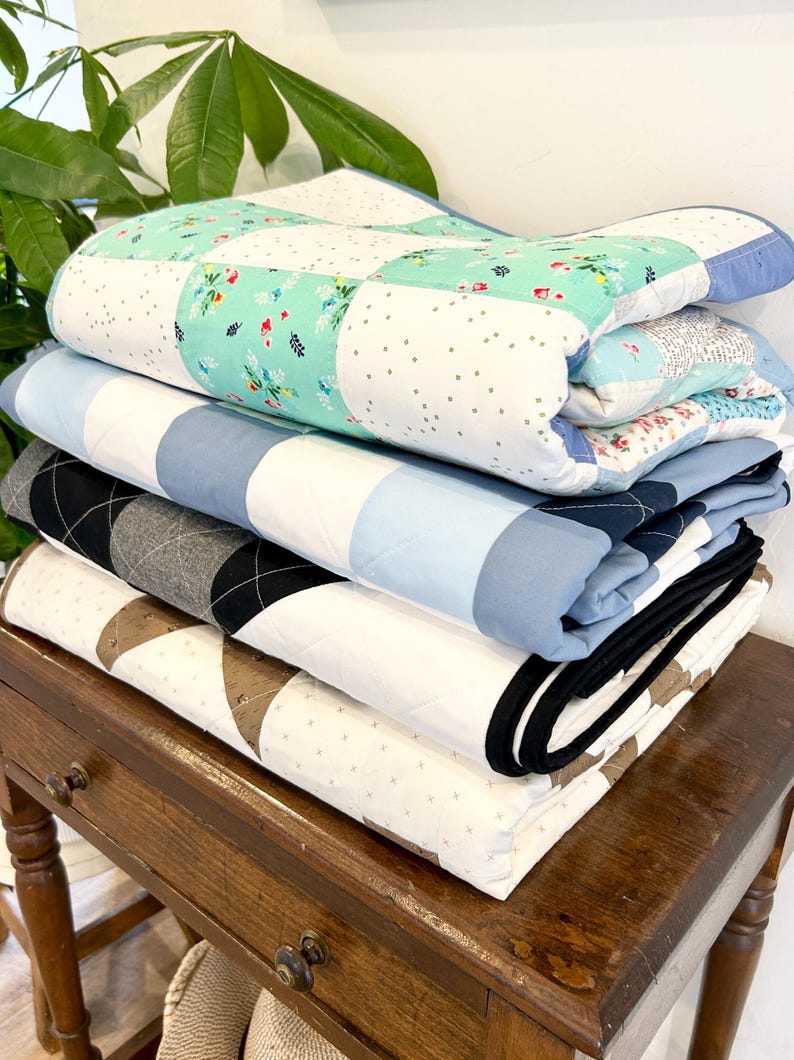 K&ouml;nnte beinhalten: Ein Stapel von vier gefalteten Quilts in verschiedenen Mustern und Farben, darunter Blau, Wei&szlig;, Gr&uuml;n und Schwarz, auf einem Holztisch mit einer Schublade.