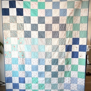 K&ouml;nnte beinhalten: Eine Patchwork-Decke mit einem Schachbrettmuster in Blaut&ouml;nen, Gr&uuml;n und Wei&szlig;. Die Decke besteht aus vielen verschiedenen Stoffquadraten, jedes mit einem einzigartigen Muster.