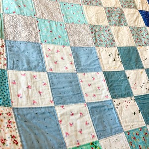 K&ouml;nnte beinhalten: Eine Patchwork-Decke mit einem karierten Muster in Blaut&ouml;nen, Wei&szlig; und Gr&uuml;n. Die Decke besteht aus verschiedenen Stoffen mit unterschiedlichen Mustern, darunter Blumenmuster, Punkte und einfarbige Stoffe.